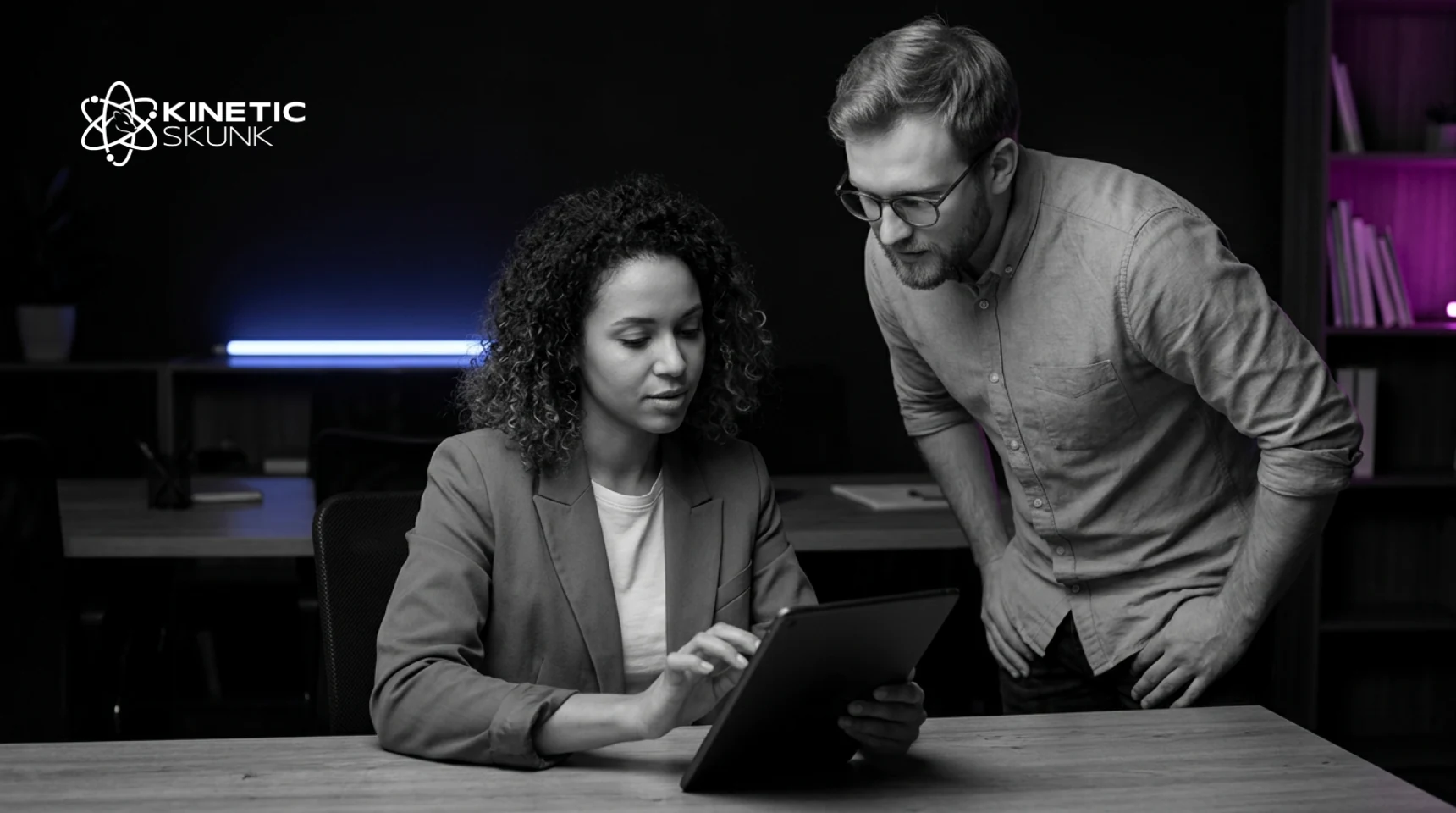 A seated colleague works through a task on a tablet while a standing colleague leans in to watch