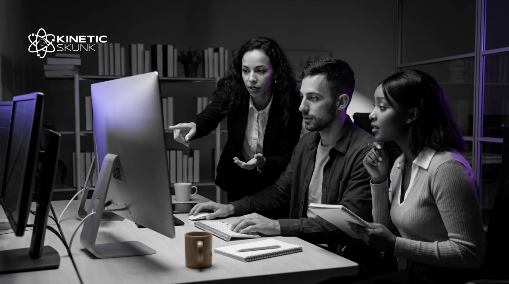 A colleague points at a monitor while an engineer at the keyboard and a teammate with a notebook follow along