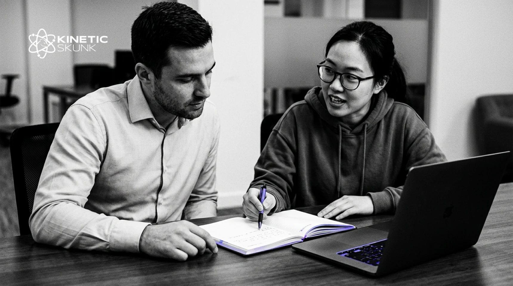 Two colleagues at a table beside a laptop, one writing in a notebook as she talks the other through her notes